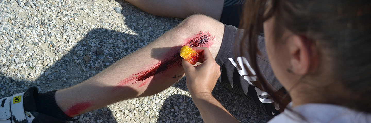 Bei einem Training zur Notfalldarstellung trägt eine Person mit einem Schwamm Kunstblut auf das Knie einer anderen Person auf und schminken fachmännisch eine realistische Wunde, während beide auf einer Kiesfläche sitzen.