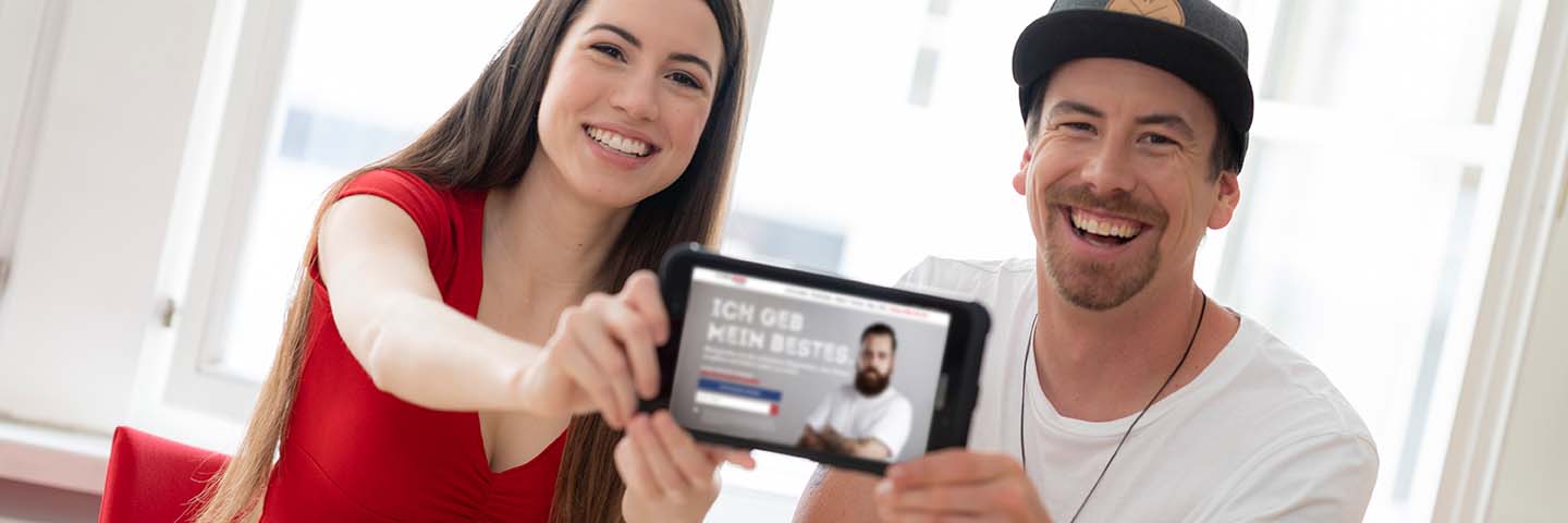 Zwei lächelnde Menschen sitzen in einem Haus und halten ein Tablet in die Kamera, auf dem eine Website über eine Blutspendeaktion mit einem bärtigen Mann und dem deutschen Text "ICH GEB MEIN BESTES" zu sehen ist. Die hell beleuchtete Szene unterstreicht die Bedeutung der Blutspende.