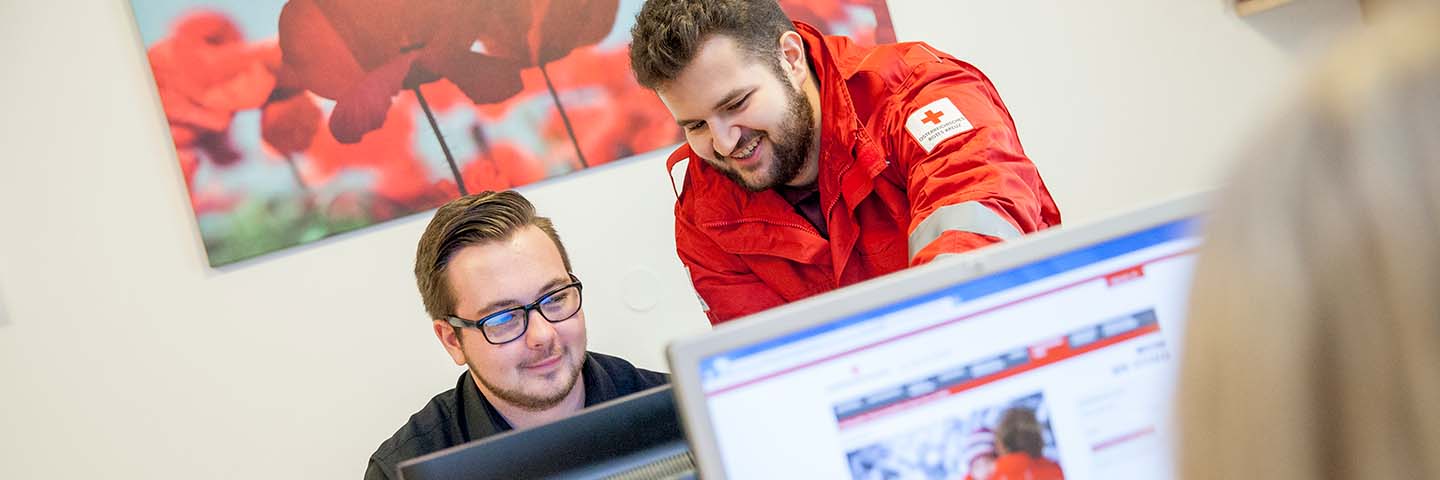 Zwei Menschen arbeiten an einem Computer; einer sitzt und trägt eine Brille, während der andere, in einer roten Jacke mit dem Emblem des Roten Kreuzes, daneben steht und lächelnd auf den Monitor zeigt - eine Szene, die die ehrenamtliche Tätigkeit unterstreicht. Ein Blumengemälde ziert die Wand hinter ihnen.